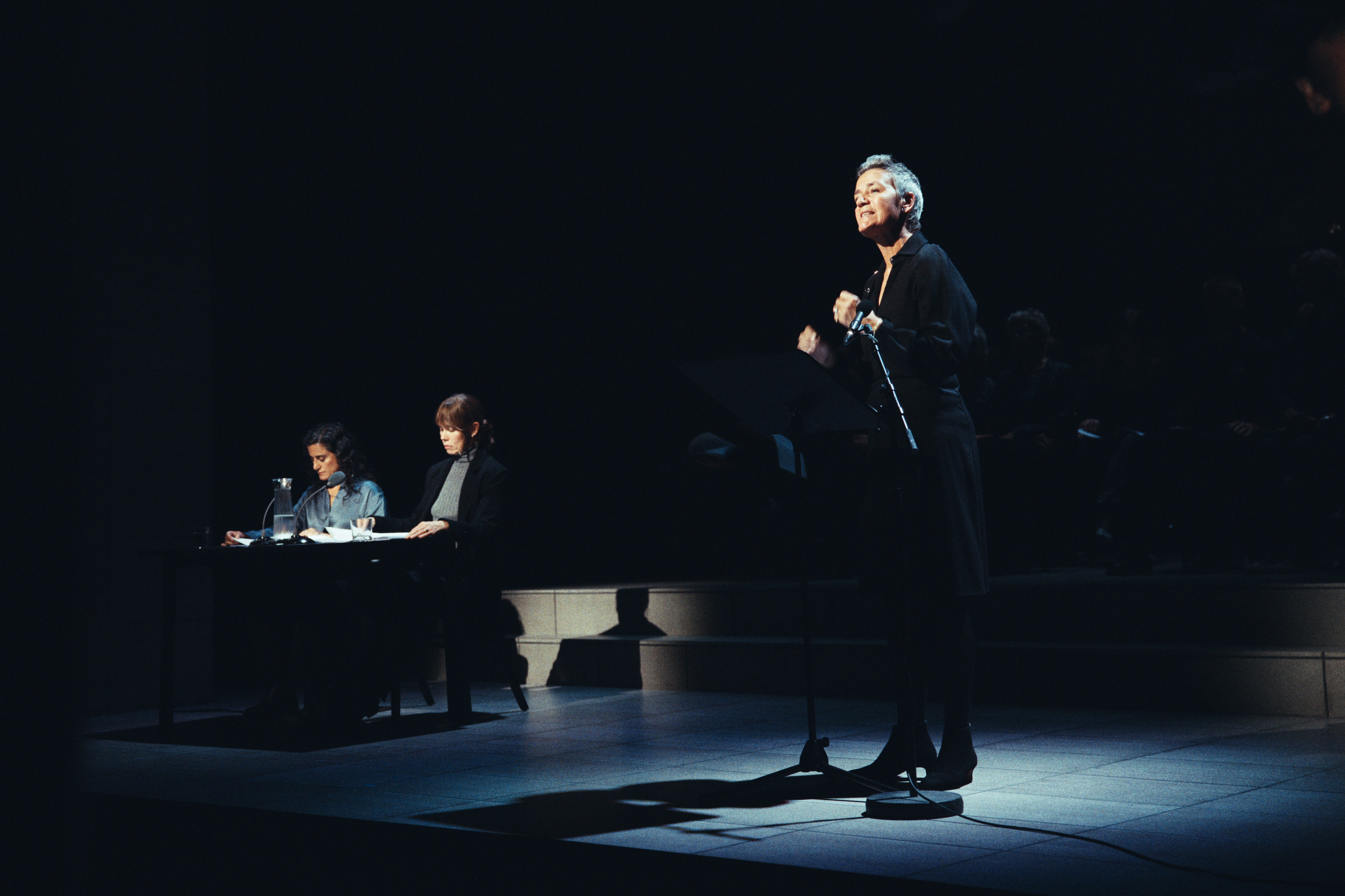 A reader standing at a microphone in the Copenhagen production of The Pelicot Trial, with two readers seated beside scripts at a table.
